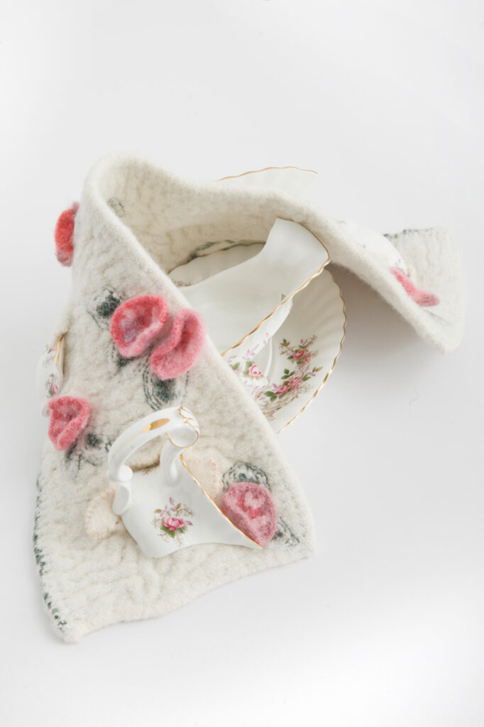 broken white china gravy jug dwcorated with pink roses wrapped in aknitted blanket, with broken pieces embedded, covered in crochet roses.